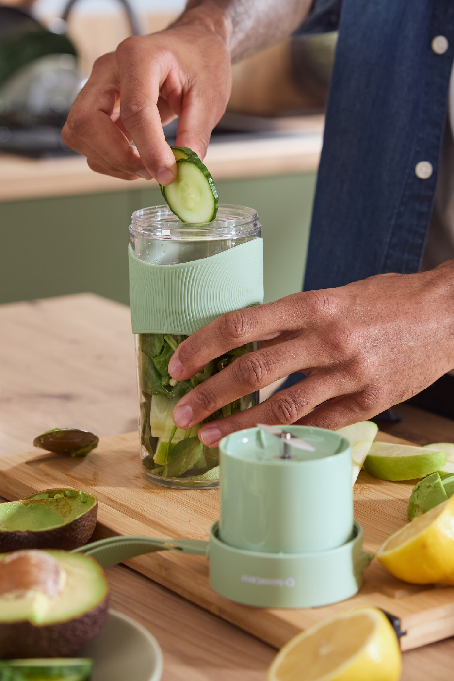 Άτομο προσθέτει αγγούρι σε φορητό μπλέντερ με σπανάκι, αβοκάντο και λεμόνι σε ξύλινη σανίδα.