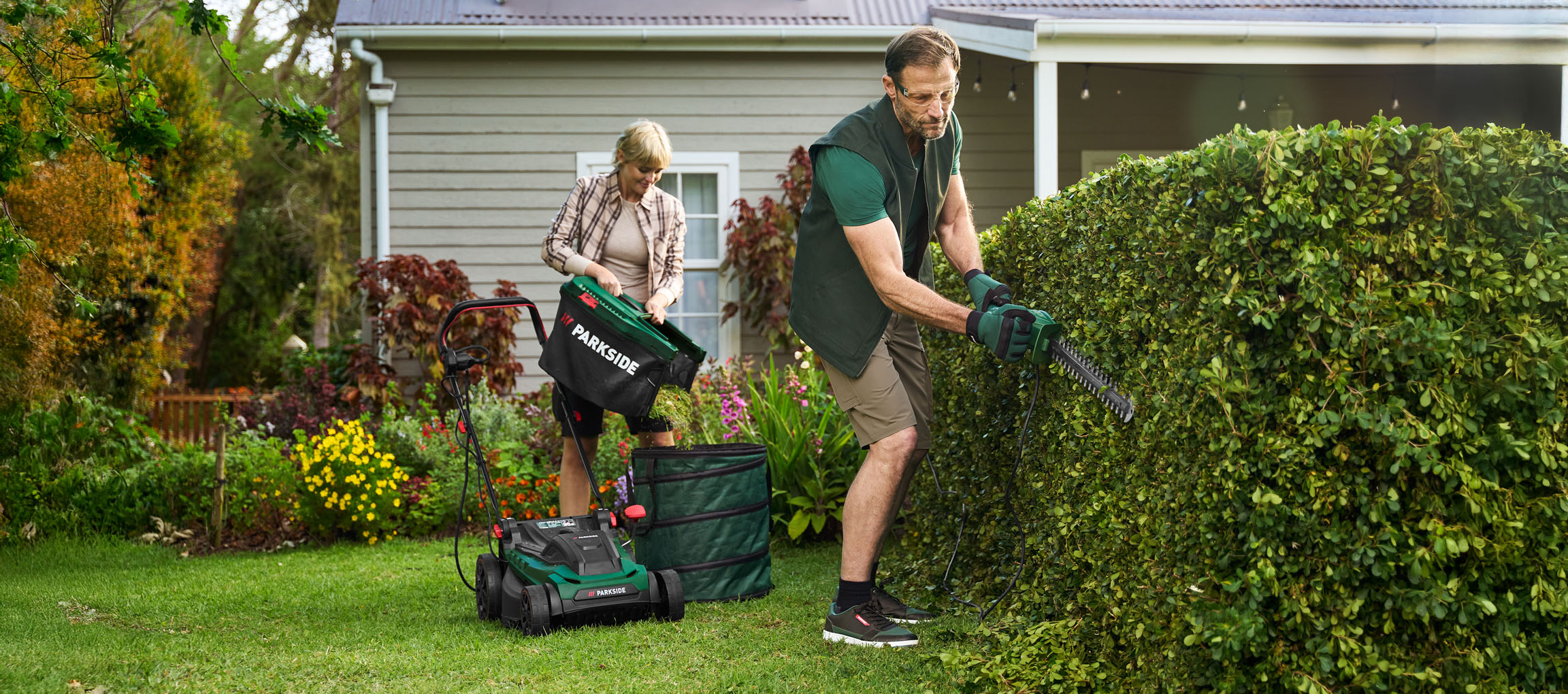 Man trims hedge, woman empties lawnmower in garden.