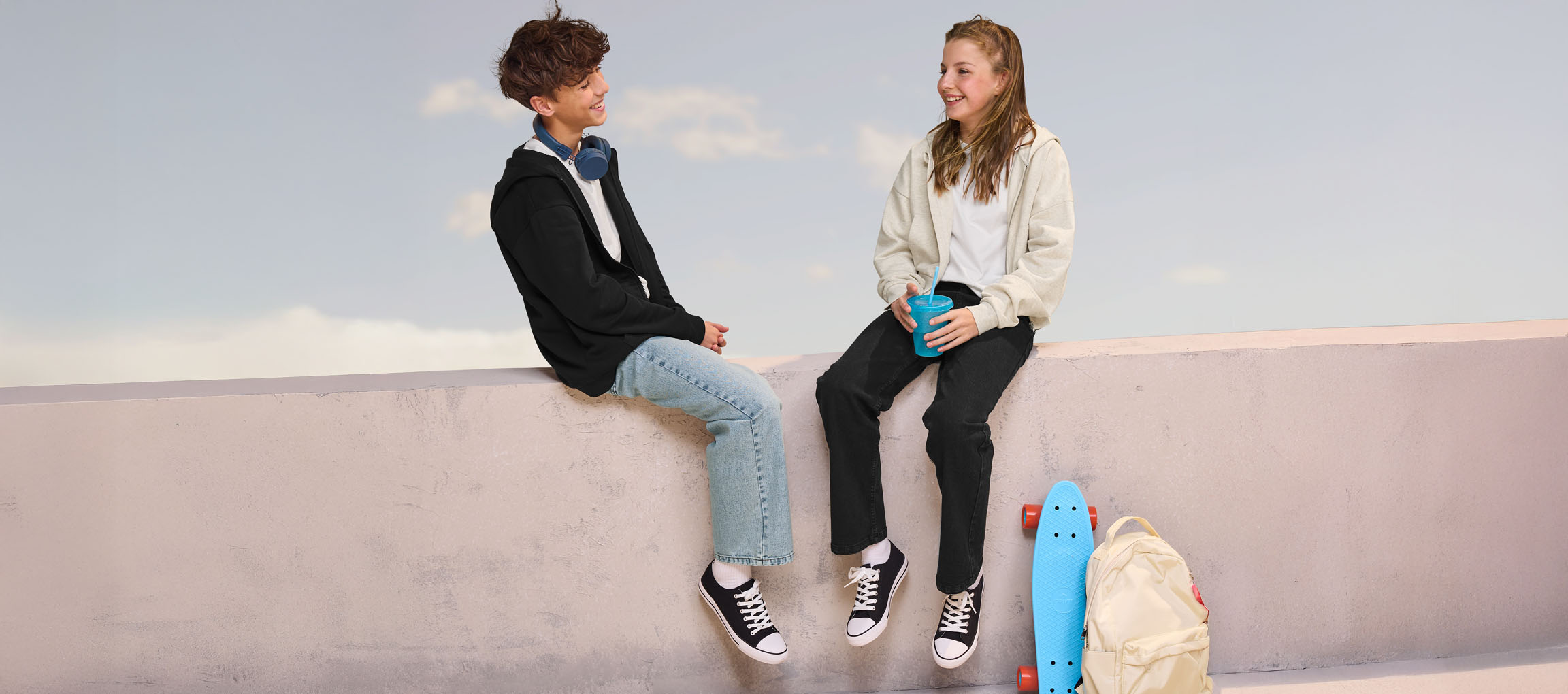 Two teenagers in hoodies and jeans sitting on a wall, with a skateboard and backpack nearby.