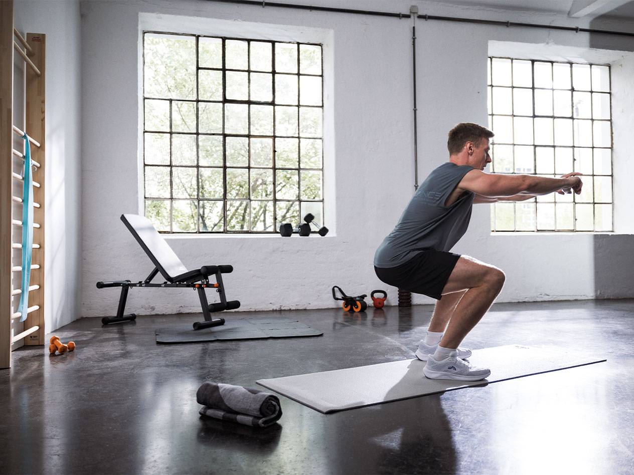 Man doing squats in a gym with a weight bench and mats.