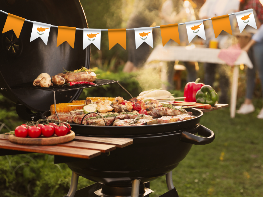 A barbecue grill filled with meat and vegetables sits outdoors with a Cyprus flag bunting in the background.