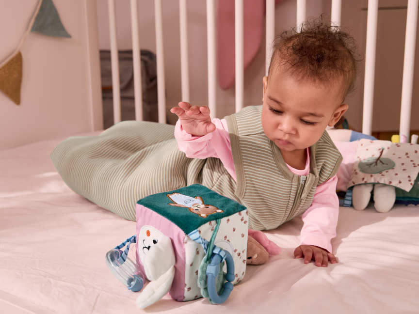A baby in a sleep sack plays with a soft activity cube in a crib.