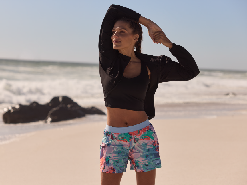 Woman in floral print shorts and black jacket stretches on the beach.