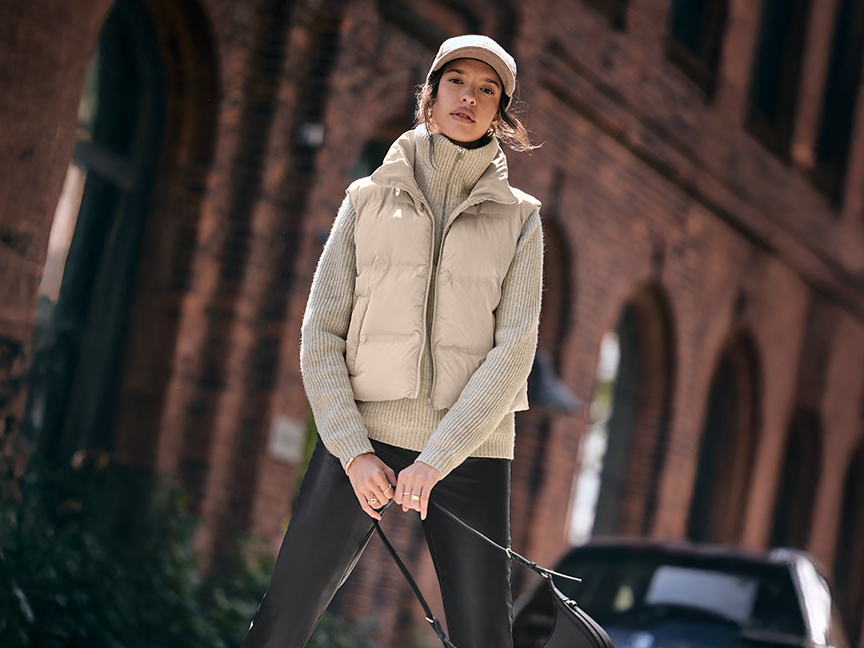 A woman in a beige puffer vest, light green turtleneck, black pants, and a cap.