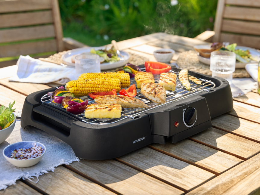 A SilverCrest electric grill cooks chicken and vegetables outdoors on a wooden table.