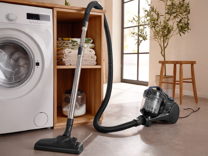 A vacuum cleaner next to a washing machine and wooden shelves with folded laundry.