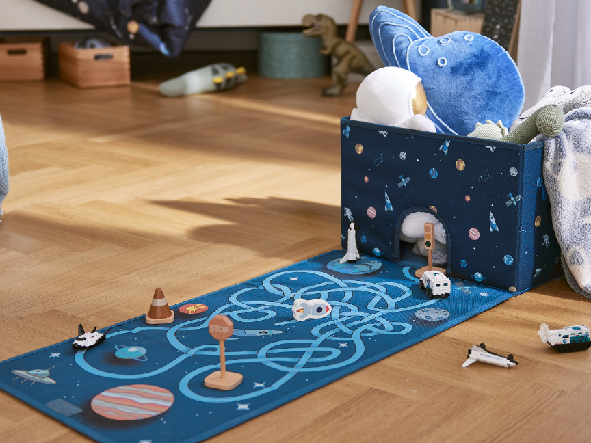 A space-themed playmat with a storage box, toys, and a blanket on a wooden floor.