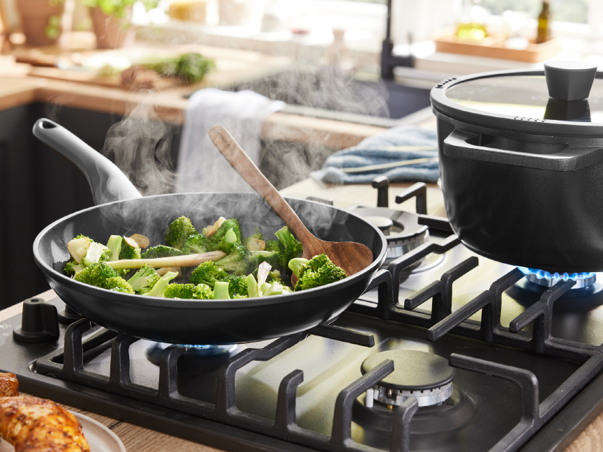 A black frying pan with steaming broccoli and a pot on a gas stove in a kitchen.