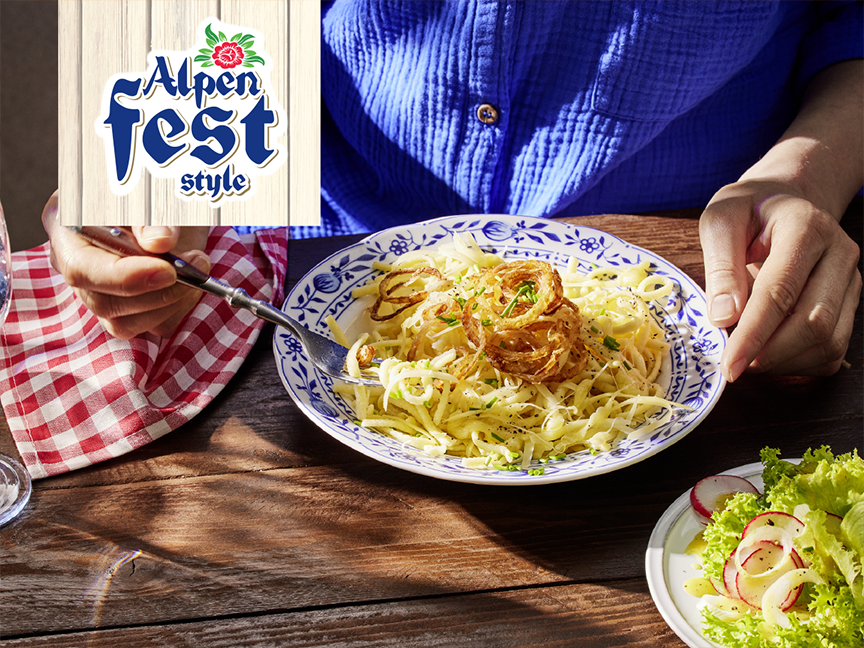 Person eating a plate of pasta with fried onions, with a salad and Alpen Fest logo.