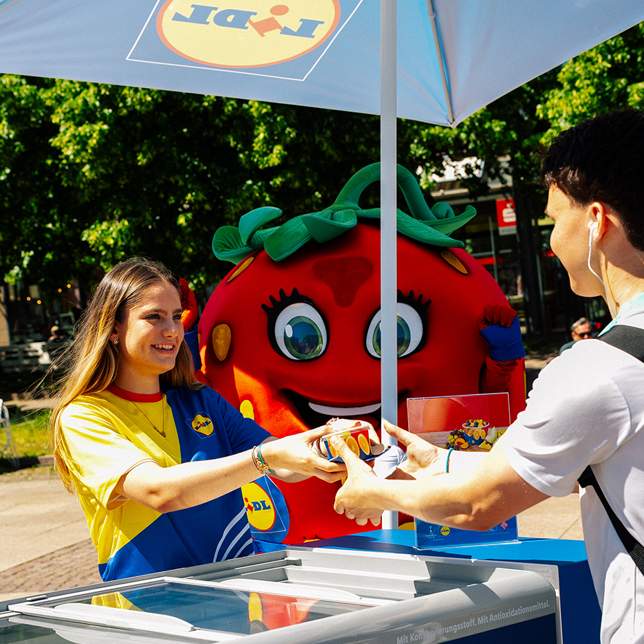 Žena daje zamrznuti desert muškarcu na Lidl štandu, a pored nje stoji i maskota jagode.