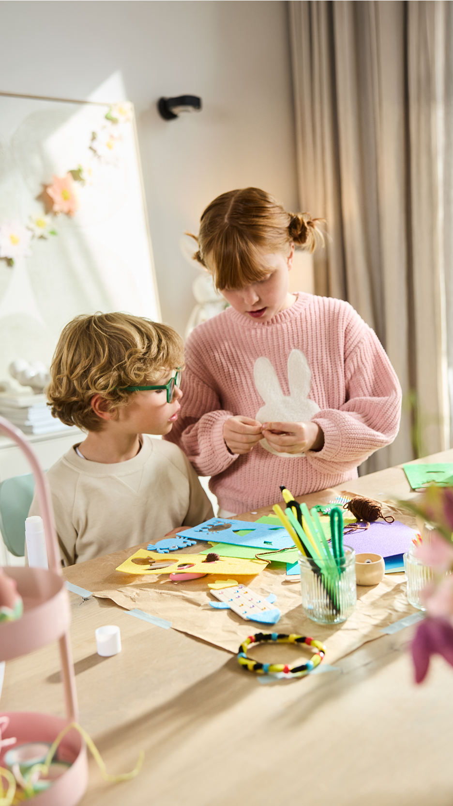 Δύο παιδιά φτιάχνουν πασχαλινές διακοσμήσεις, συμπεριλαμβανομένου ενός κουνελιού, με διάφορα υλικά χειροτεχνίας.