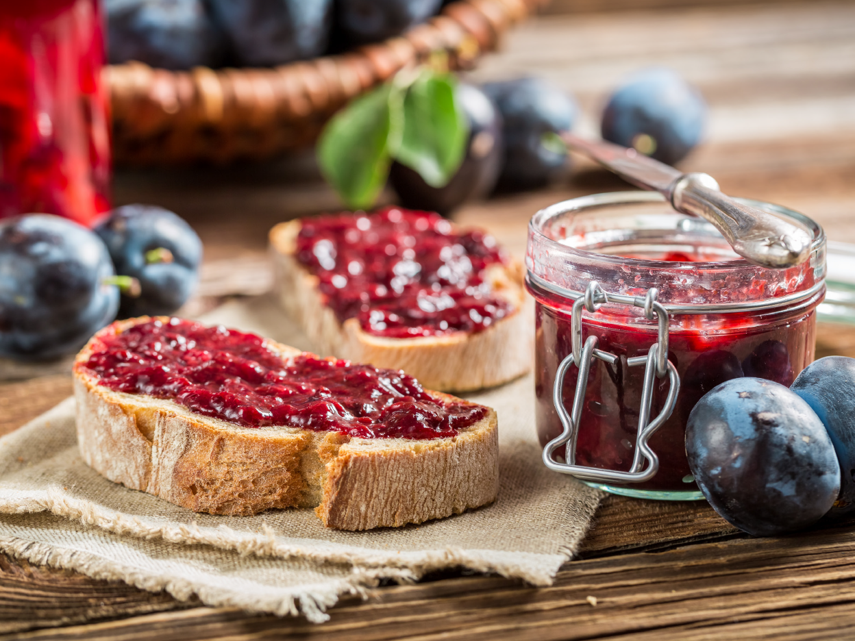 Plum jam on bread slices, with a jar of jam and fresh plums on a wooden table.