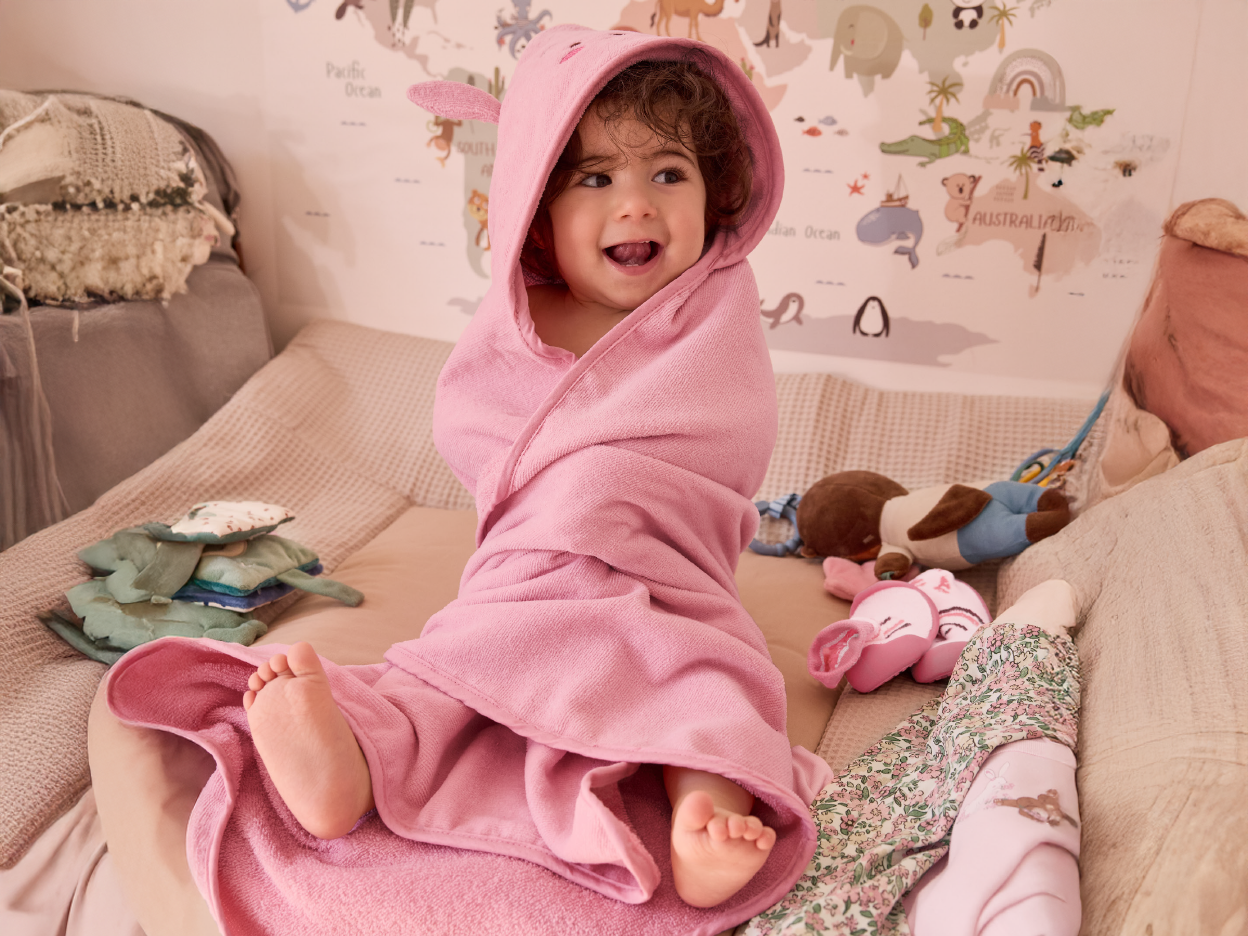 A baby wrapped in a pink hooded towel, sitting on a bed with toys.