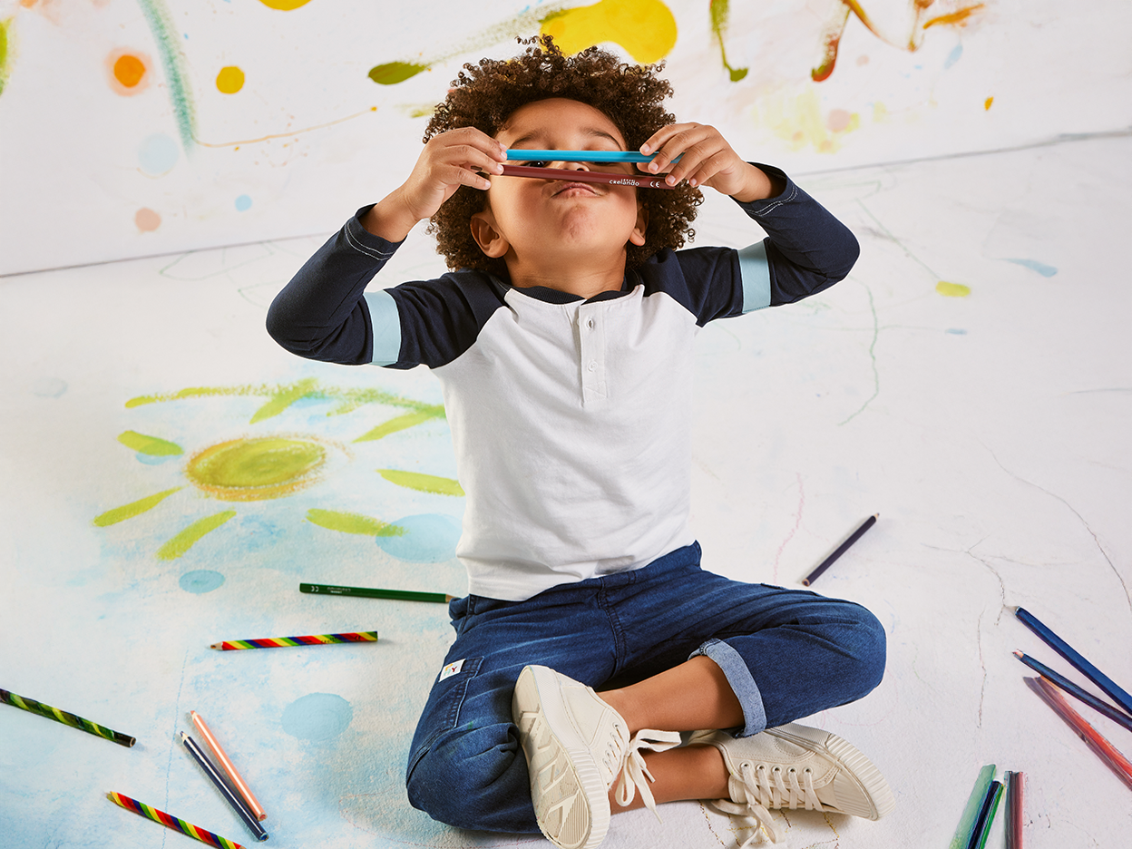 A young boy holds two colored pencils to his eyes, sitting on a floor with drawings.