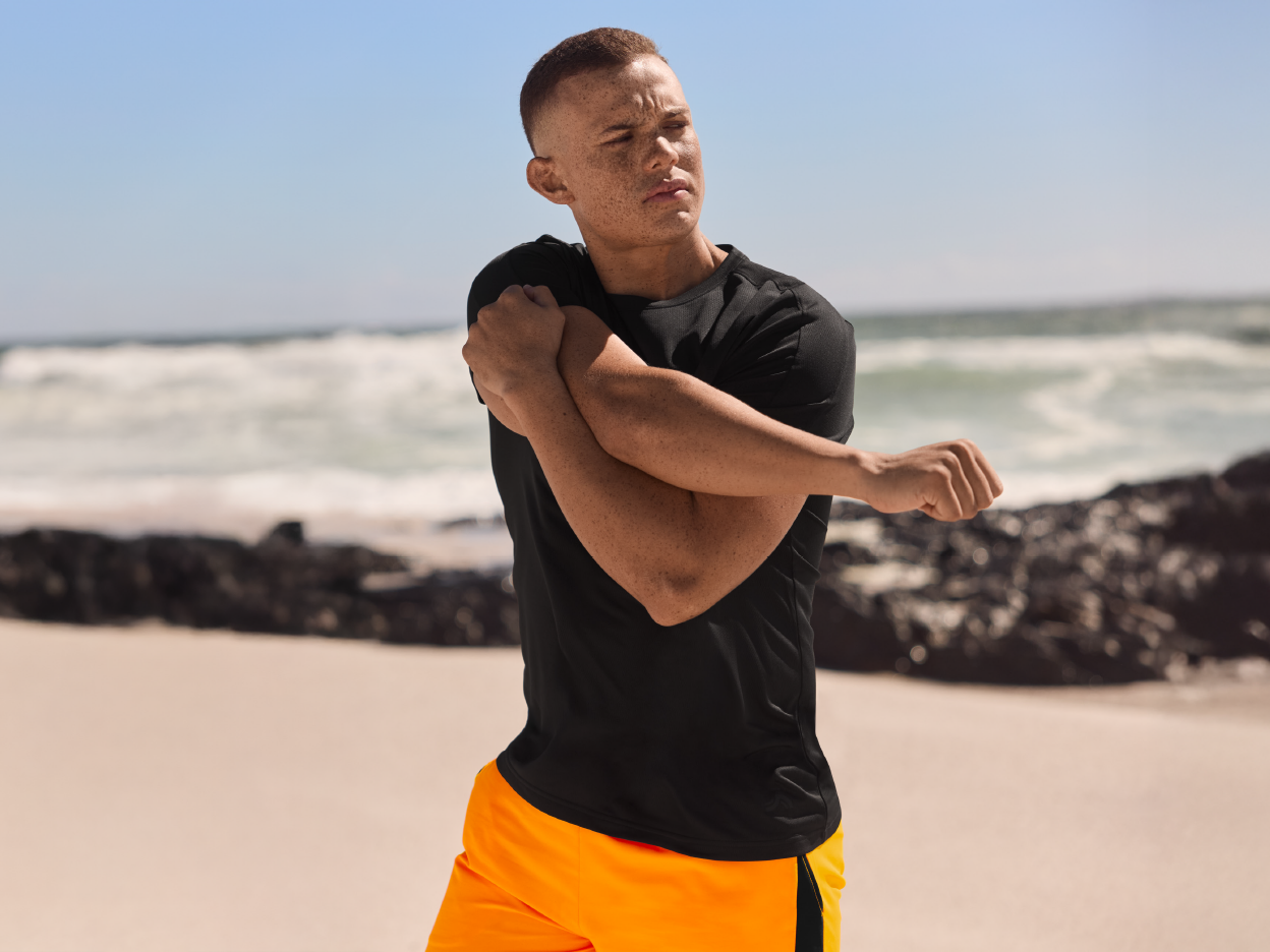 A man stretches his arms on a beach wearing a black shirt and orange shorts.