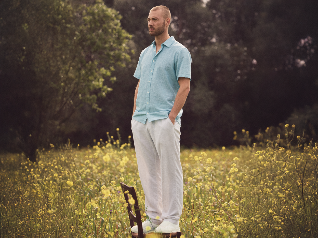 A man in a light blue linen shirt and white pants stands in a field of yellow flowers.