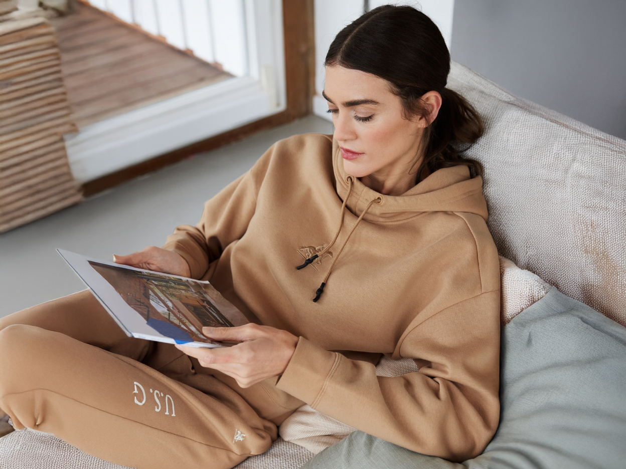 A woman in a tan hoodie and sweatpants reads a magazine on a couch.