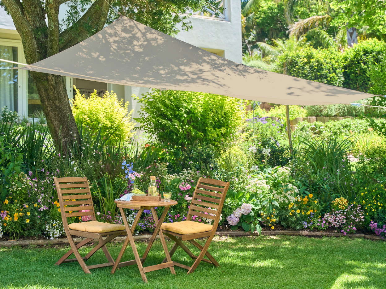 A beige triangular shade sail provides shade over a wooden patio table and chairs in a vibrant garden.