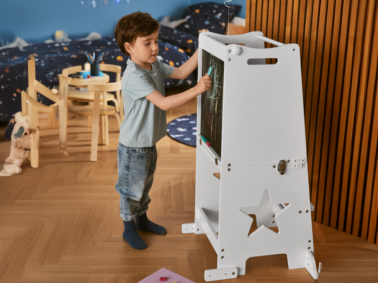 A young boy drawing on a white wooden learning tower with a chalkboard.