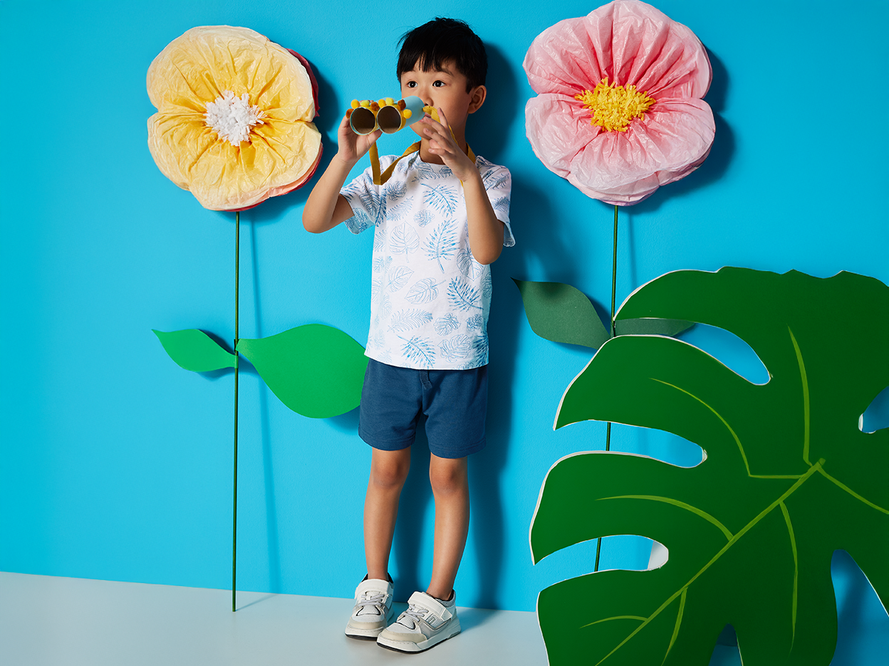 A boy in summer clothes uses cardboard binoculars to look at large paper flowers.