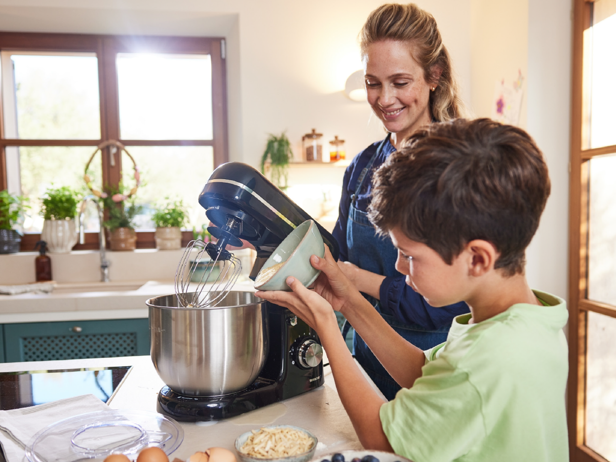 A woman and a boy baking with a black stand mixer in a bright kitchen.