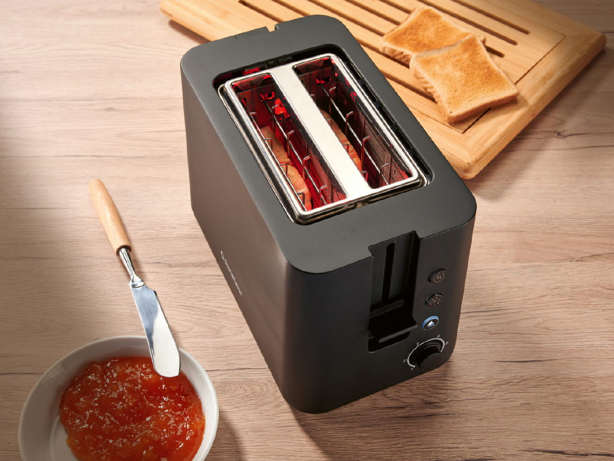 A black toaster with glowing red slots, next to a bowl of jam and toast on a wooden board.