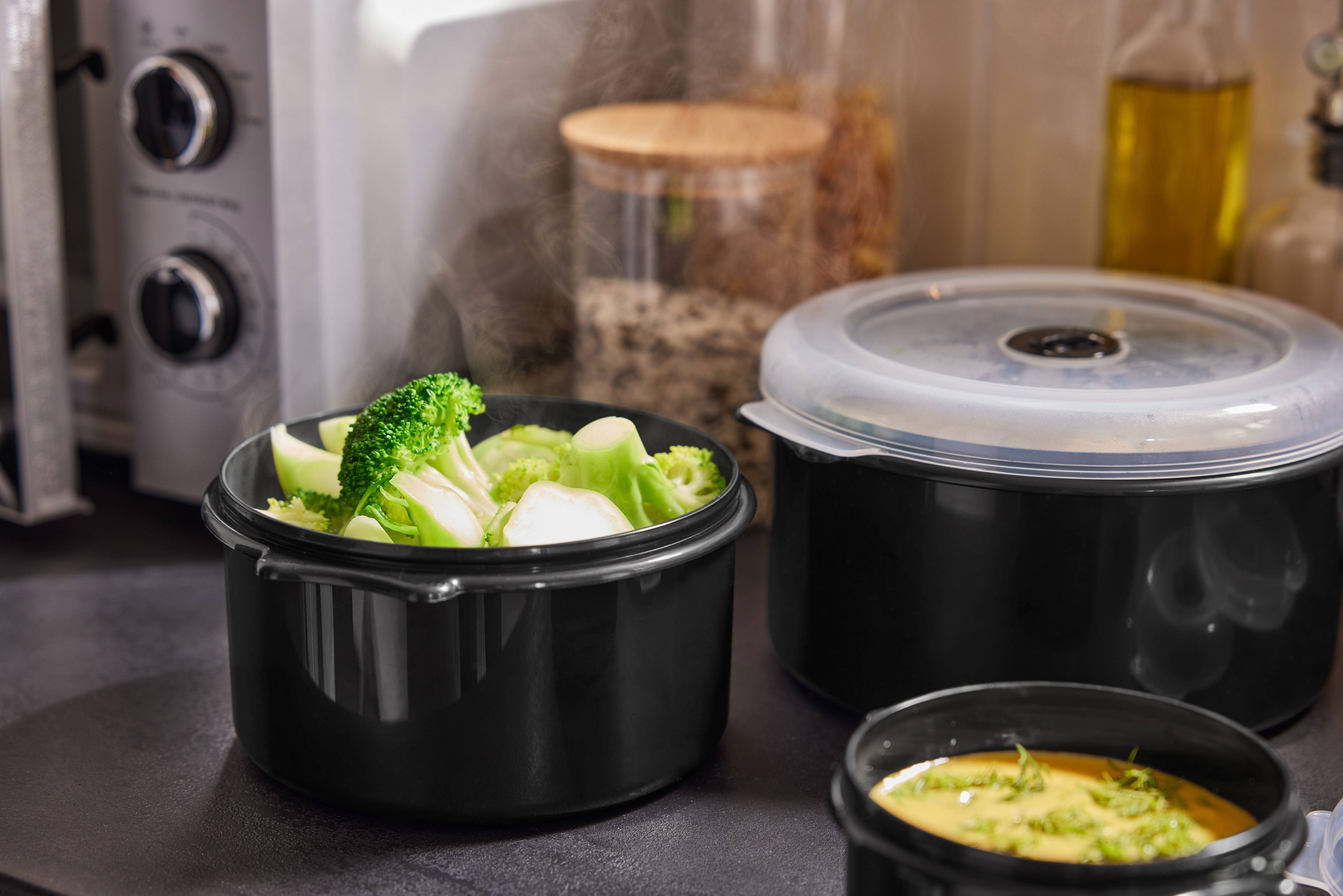 Steaming broccoli and other dishes in black containers on a kitchen counter.