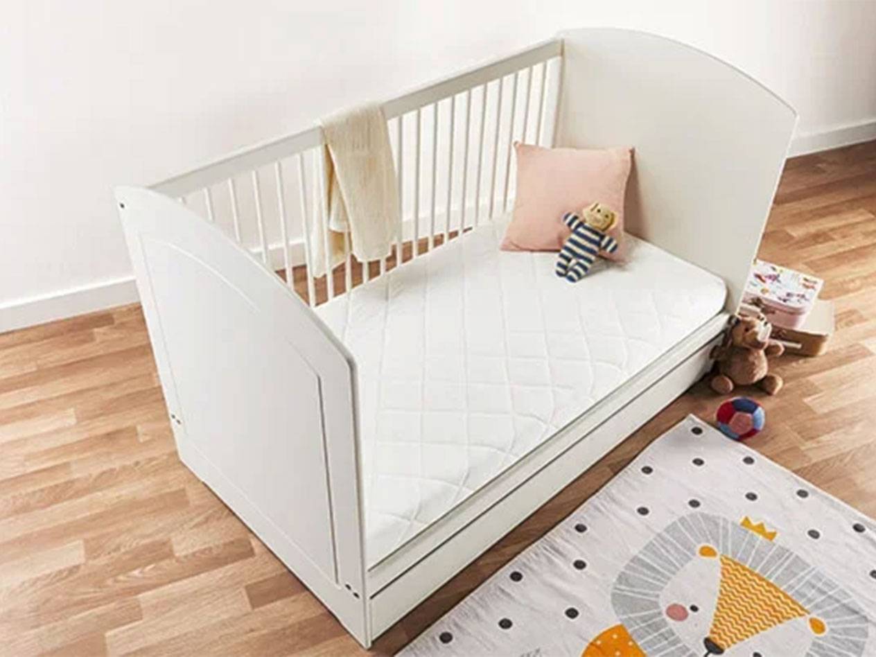 White baby cot with mattress, pillow, and toys on a wooden floor.