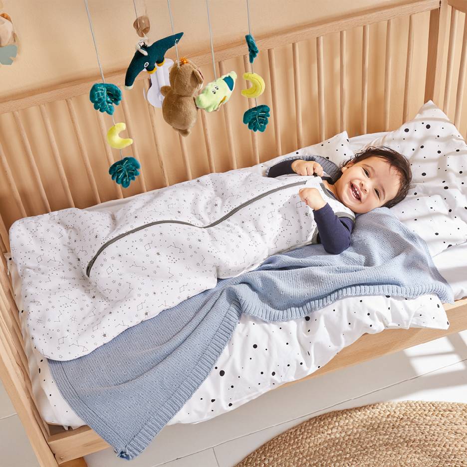 Happy baby in a star-patterned sleeping bag in a wooden crib with a toy mobile.