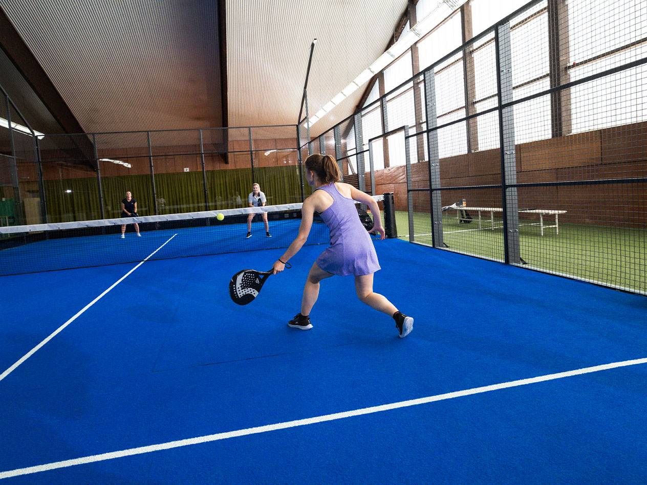 Woman playing padel on a blue court, with other players in the background.