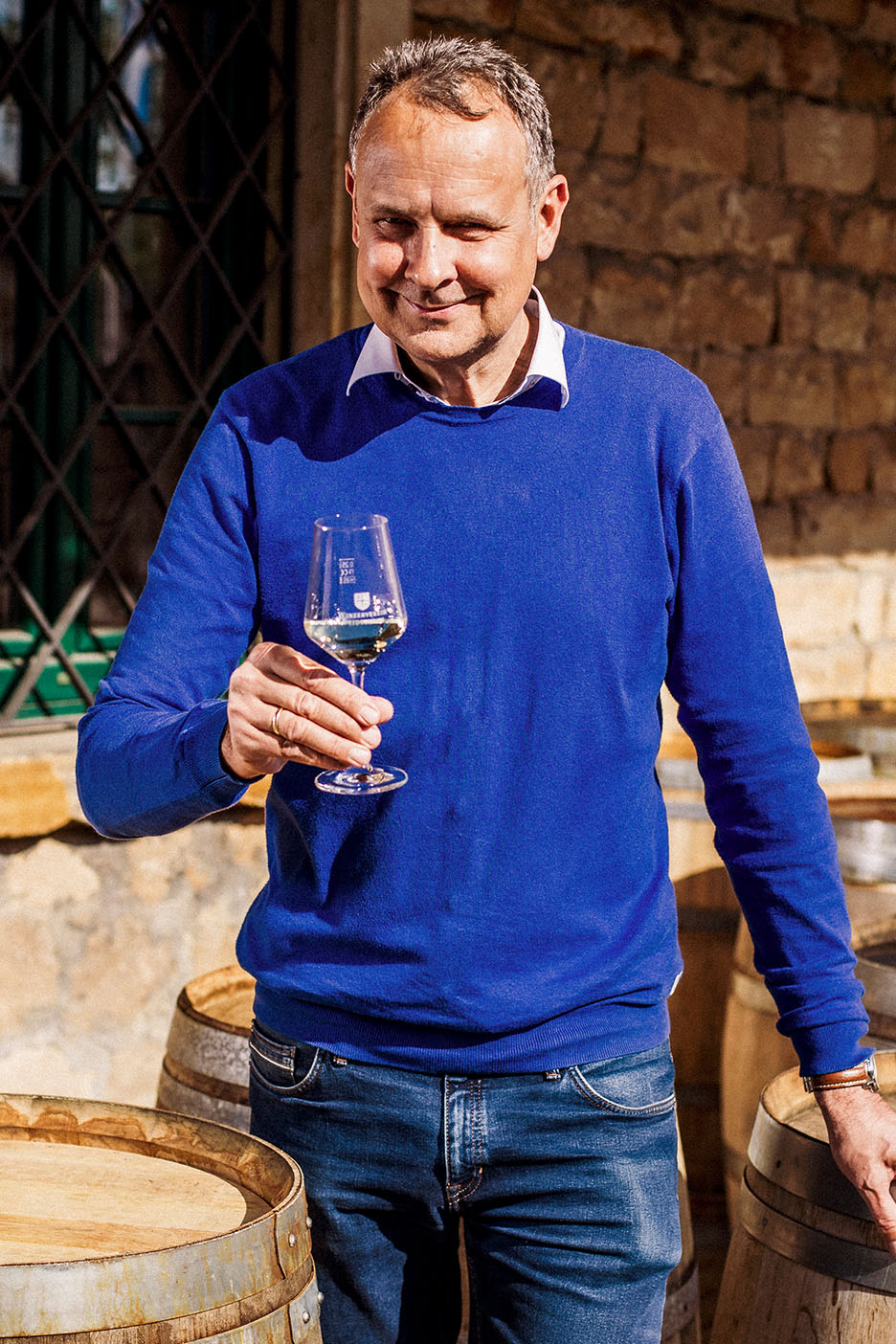 Smiling man in blue jumper and jeans, holding a glass of white wine among wooden barrels.
