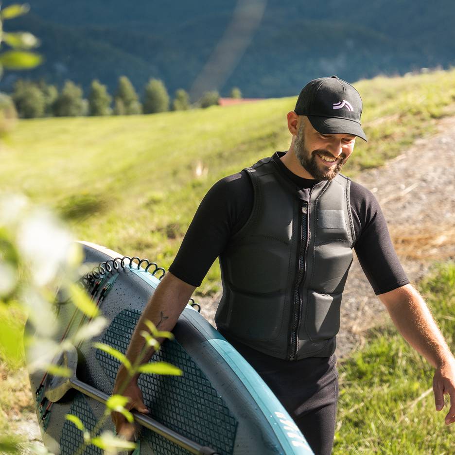Man in black wetsuit and life vest, carrying a SUP board with a paddle.