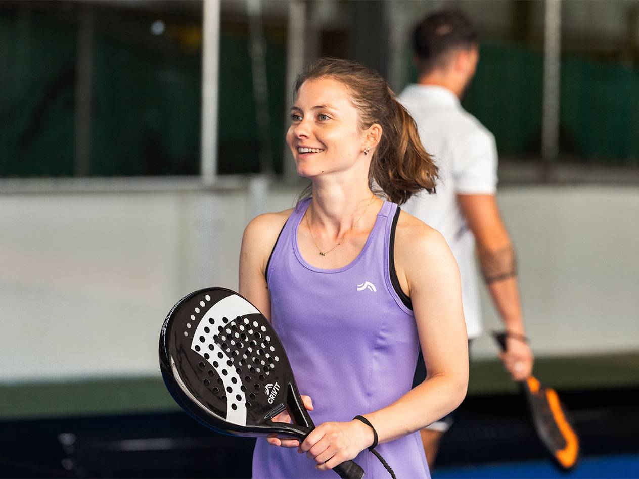 Woman in a purple sports dress holding a padel racket, man in the background.