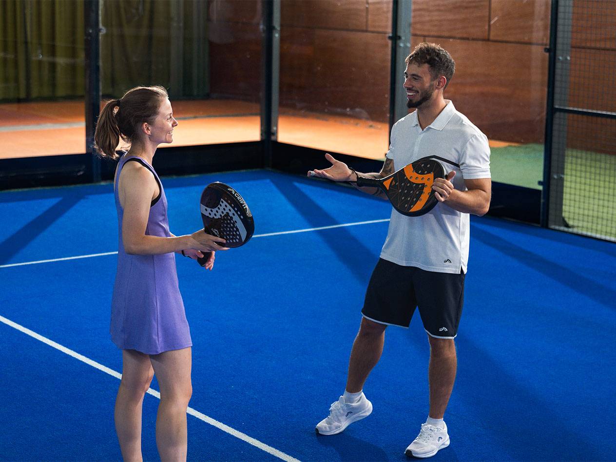 Two padel players with rackets on a blue court, smiling at each other.