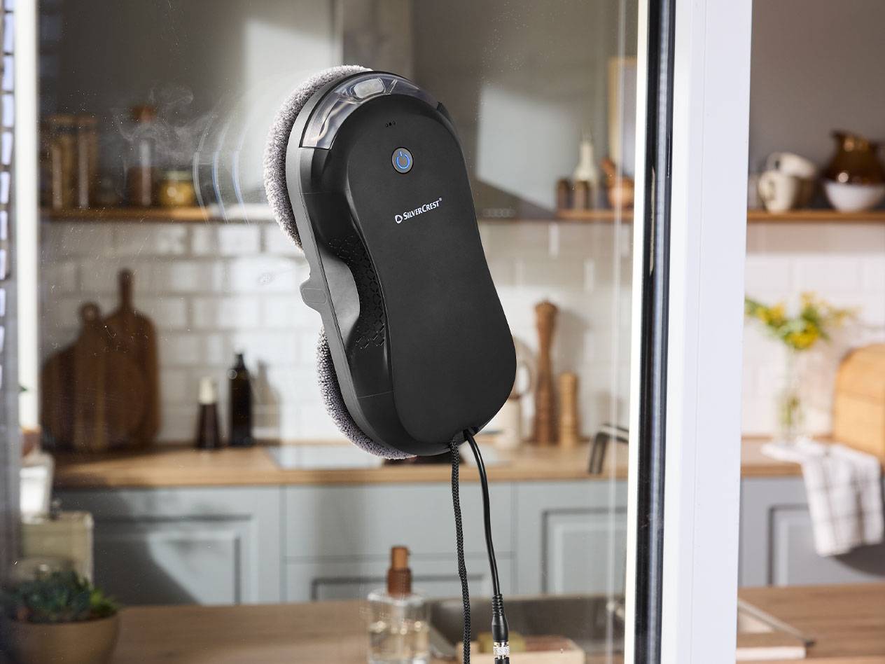 Ρομπότ καθαρισμού παραθύρων Silvercrest σε τζάμι με κουζίνα στο παρασκήνιο.