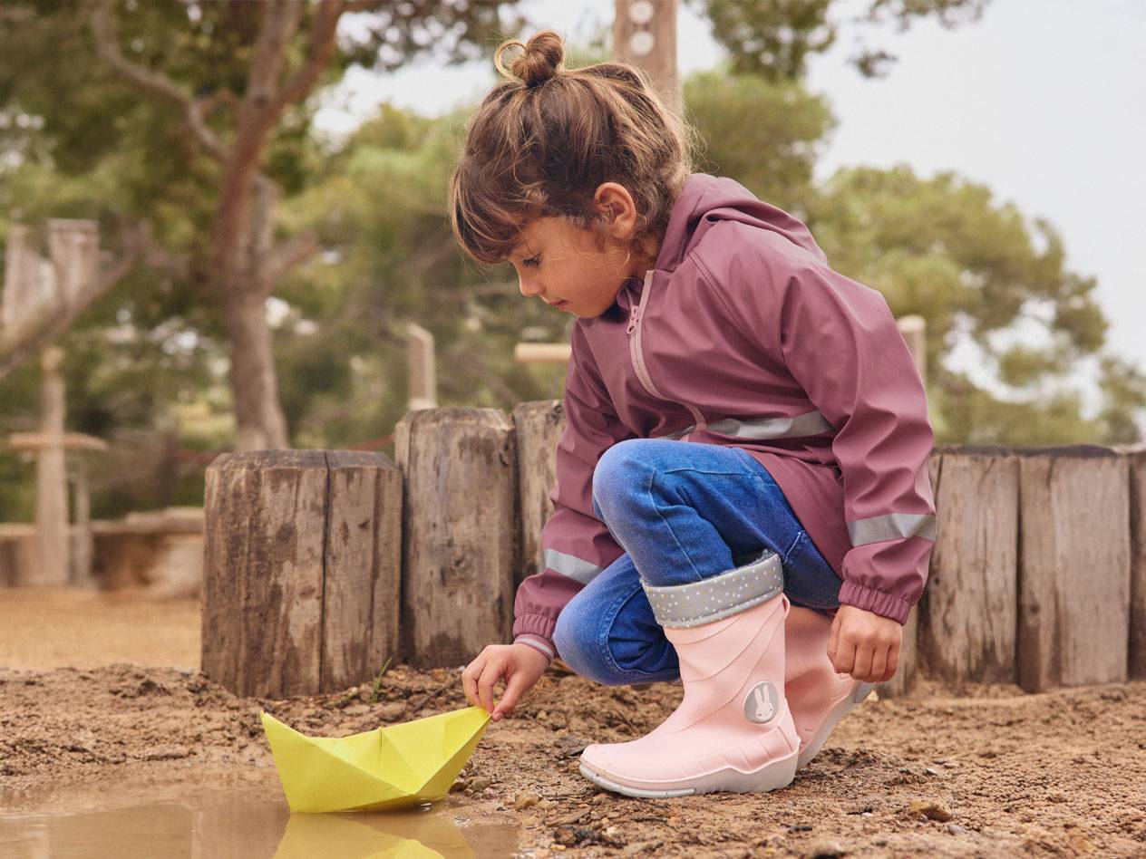 Girl in pink rain boots and jacket, playing with a paper boat in a puddle.