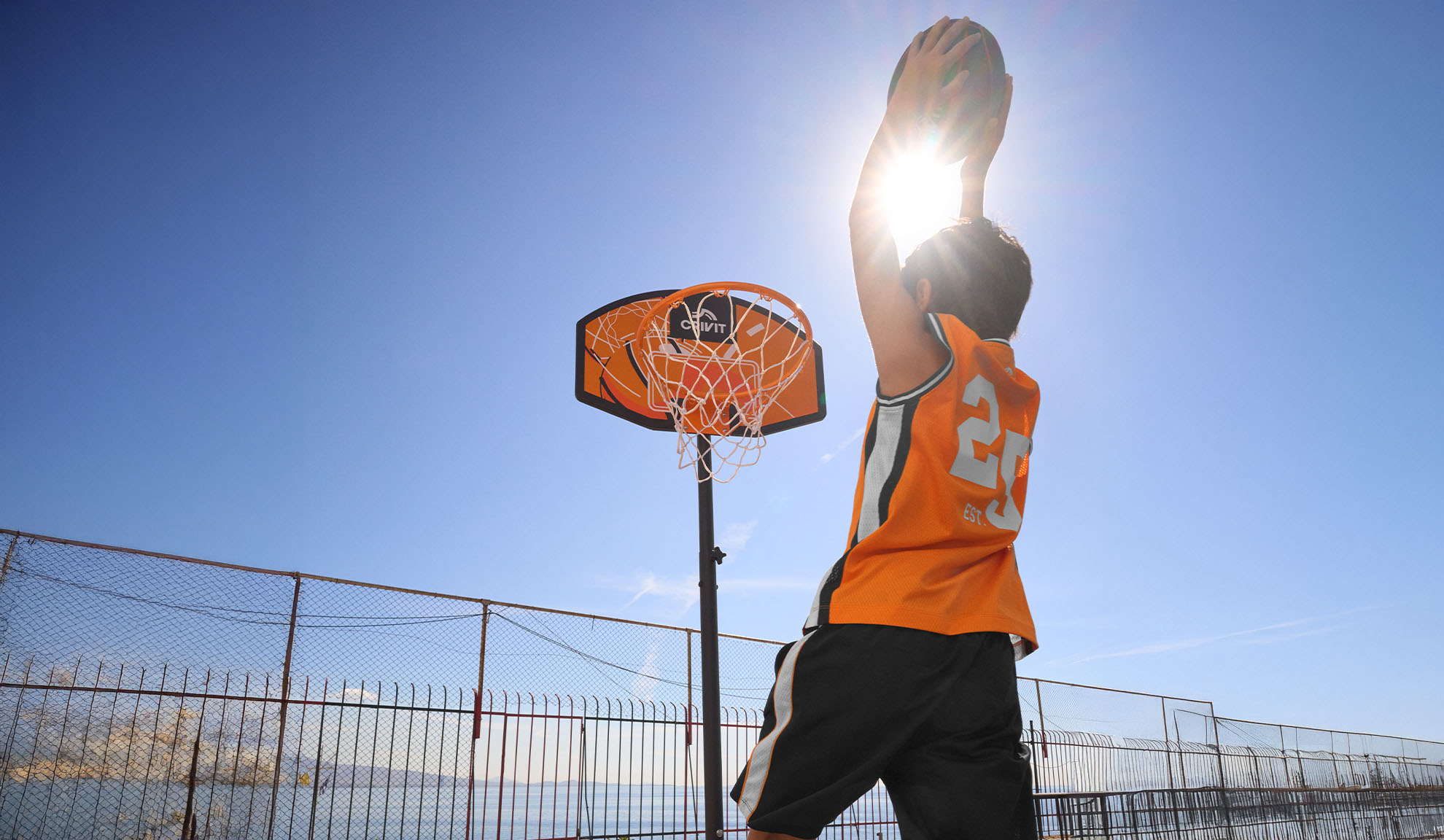 Child in basketball jersey shooting hoops outdoors with bright sun