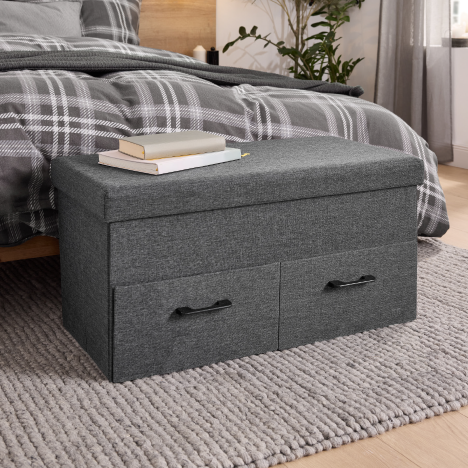 Γκρι υφασμάτινος οθωμανικός πάγκος αποθήκευσης με δύο συρτάρια στους πρόποδες ενός κρεβατιού με καρό κλινοσκεπάσματα.