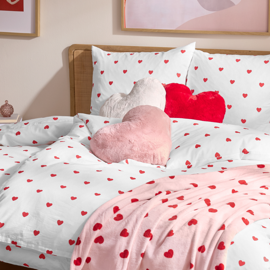 White bedding with red hearts, three heart-shaped pillows, and a pink blanket with red hearts.