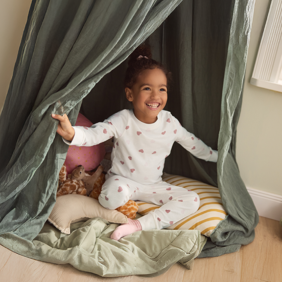 Happy child in heart-patterned pajamas sitting in a green fabric play tent with pillows.