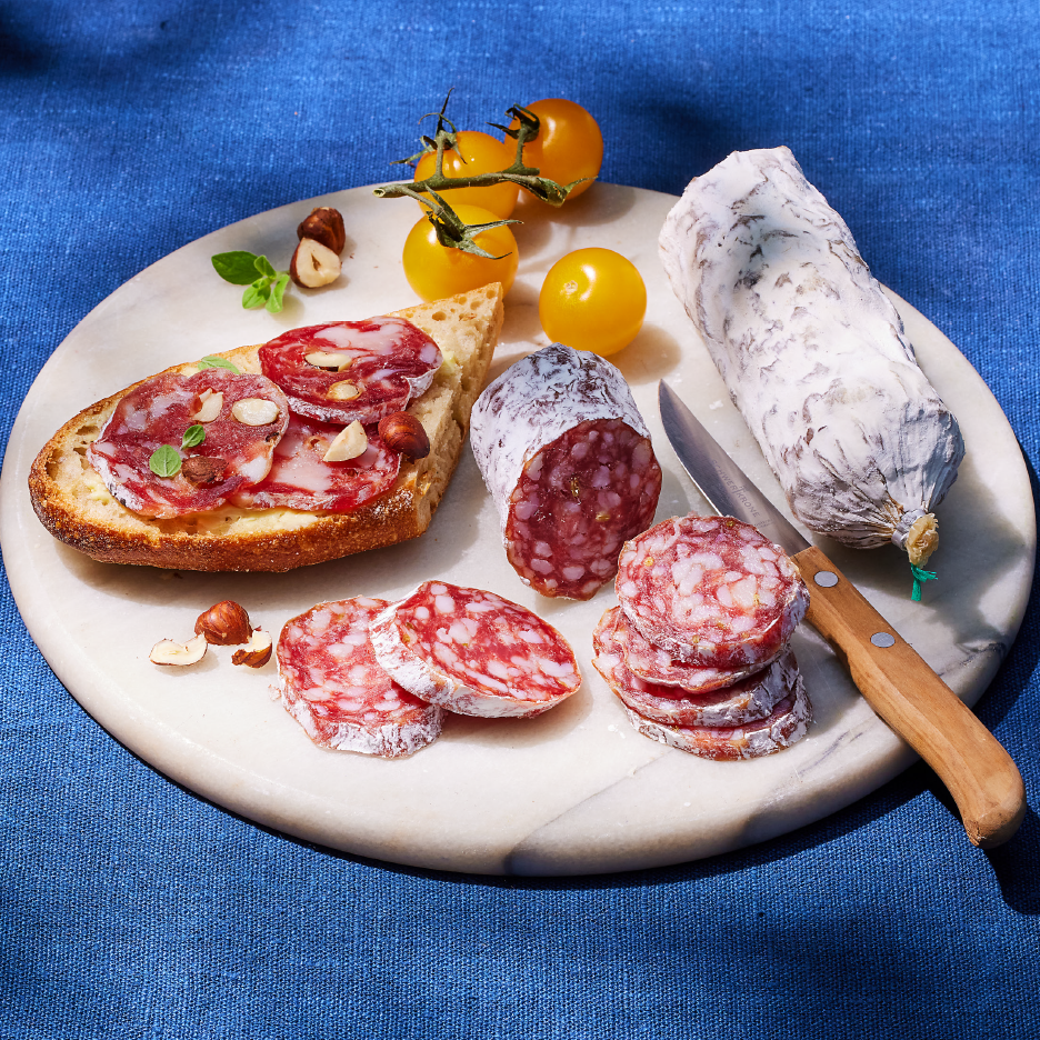 Salami slices and whole salami on a marble board with bread, hazelnuts, and tomatoes.