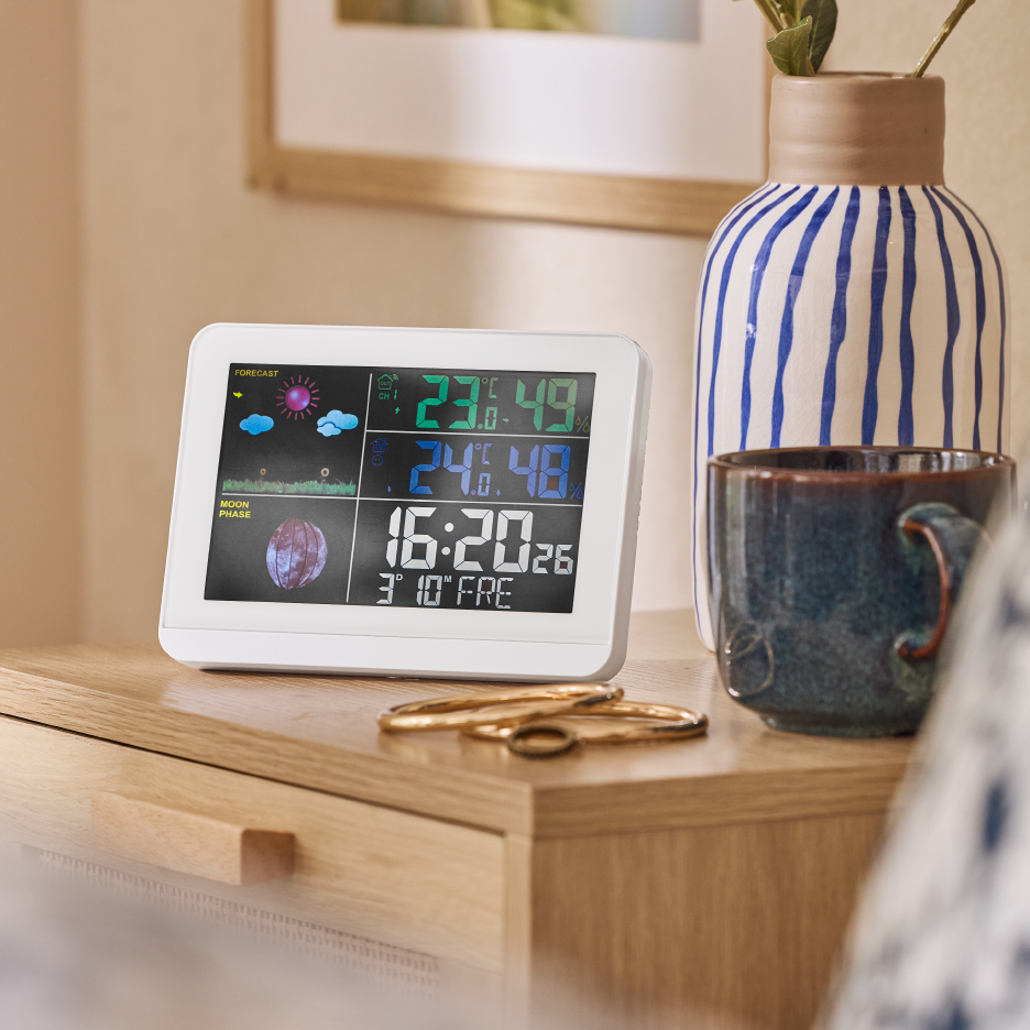 A white digital weather station displaying forecast, moon phase, temperature, humidity, and time, on a wooden bedside table next to a blue and white striped vase and a mug.