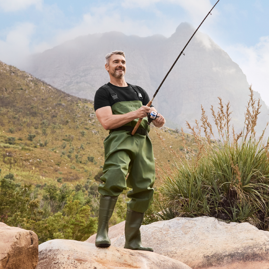 Άνδρας με αδιάβροχη στολή ψαρέματος και μπότες, κρατώντας καλάμι ψαρέματος, στέκεται σε βράχια σε εξωτερικό χώρο.
