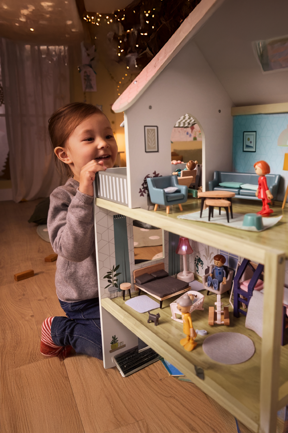 Happy child playing with a large wooden dollhouse with furniture and figures.