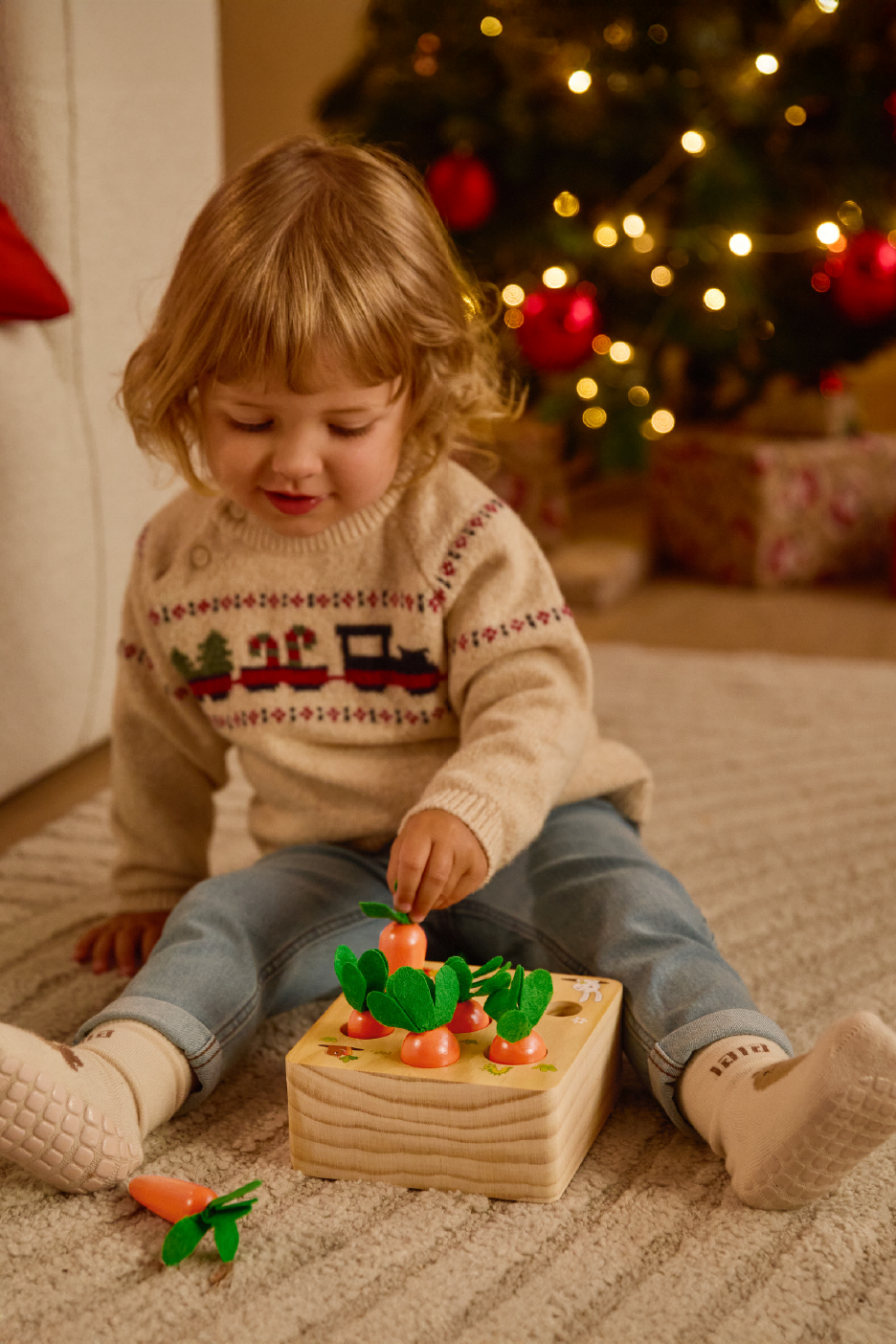 Νήπιο παίζει με ένα ξύλινο παιχνίδι καρότο, φορώντας ένα χριστουγεννιάτικο πουλόβερ.