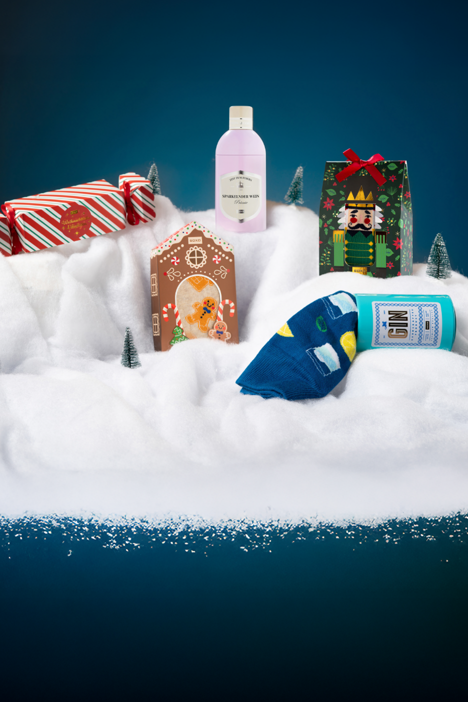 Χριστουγεννιάτικα δώρα: ροζ μπουκάλι, σπιτάκι από μελόψωμο, κουτί καρυοθραύστης, κάλτσες και κουτί τζιν στο χιόνι.