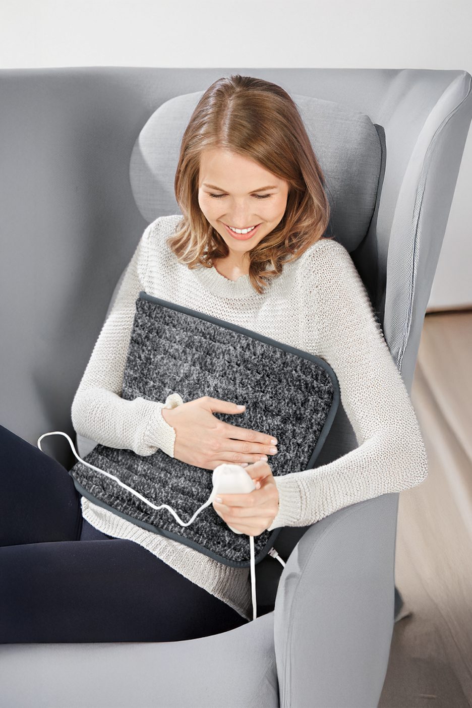 Γυναίκα σε πολυθρόνα χρησιμοποιεί μια γκρι ηλεκτρική θερμοφόρα με χειριστήριο.