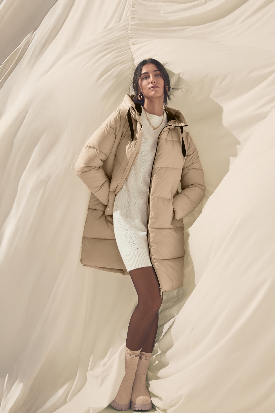 Γυναίκα με μπεζ φουσκωτό παλτό, λευκό πλεκτό φόρεμα και καφέ καλσόν με μπεζ μπότες.