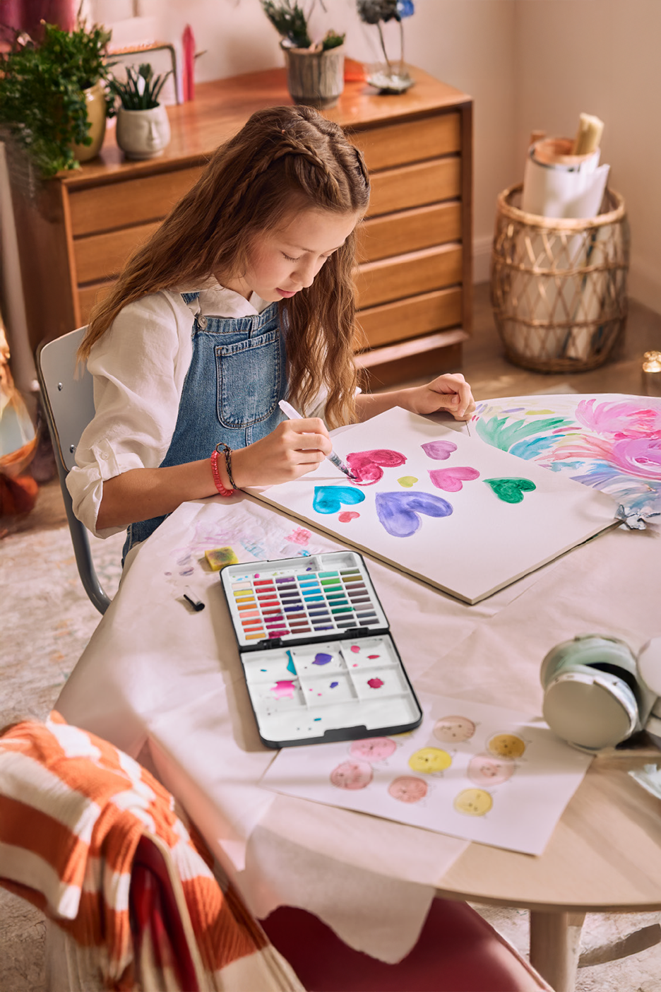 Κορίτσι ζωγραφίζει καρδιές με ακουαρέλες σε ξύλινο τραπέζι, με σετ χρωμάτων και πινέλα.