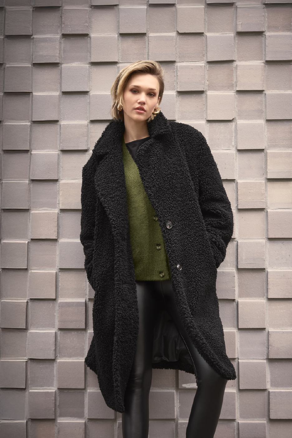 Γυναίκα με μαύρο παλτό teddy, πράσινη ζακέτα και μαύρο κολάν από ψεύτικο δέρμα.